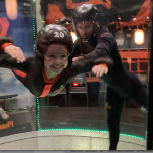 adventurous child flies in air, wearing black helmet, supported by man in black, the activity is happening in the ifly wind tube